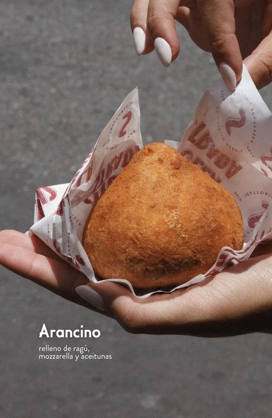 Arancino dorado apoyado sobre papel de Sulla Strada en la palma de una mano, sobre el asfalto