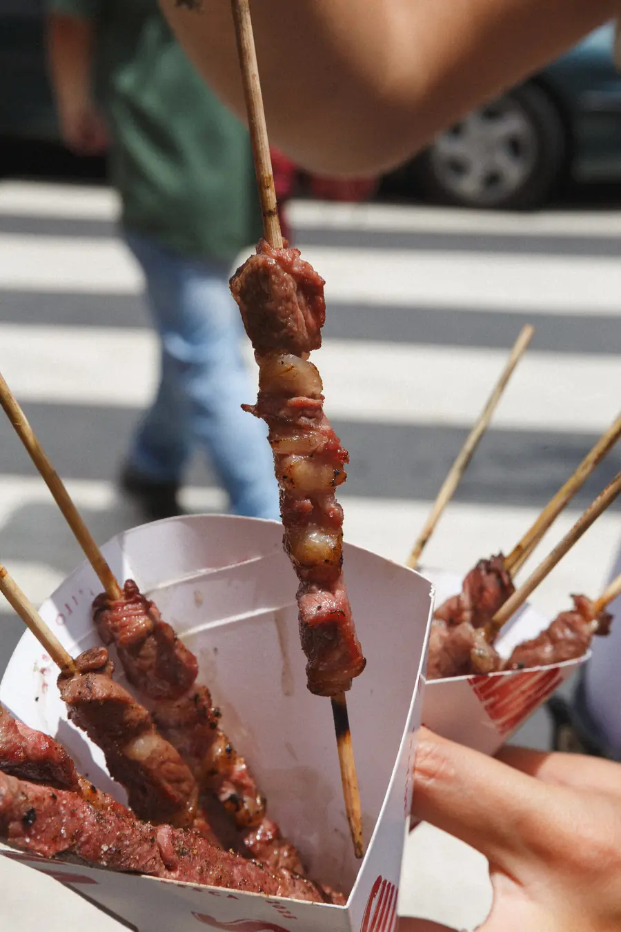 Arrosticini de carne recién asados en sus cucuruchos de papel, alzados sobre un paso de cebra