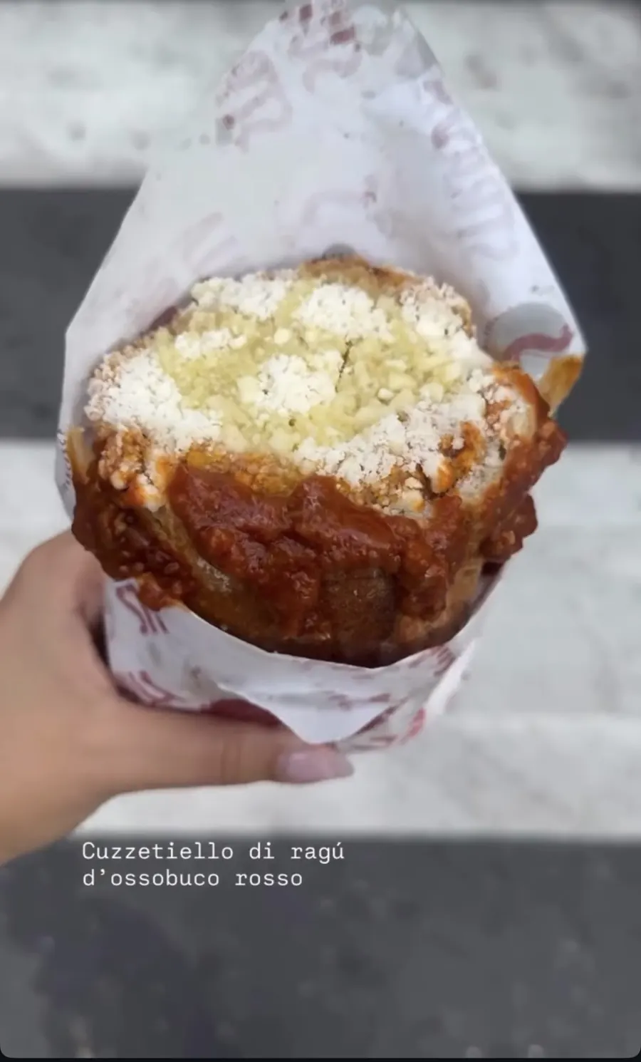 Cuzzetiello desbordado de ragú d'ossobuco rojo y queso rallado, sostenido en la calle sobre un paso de peatones