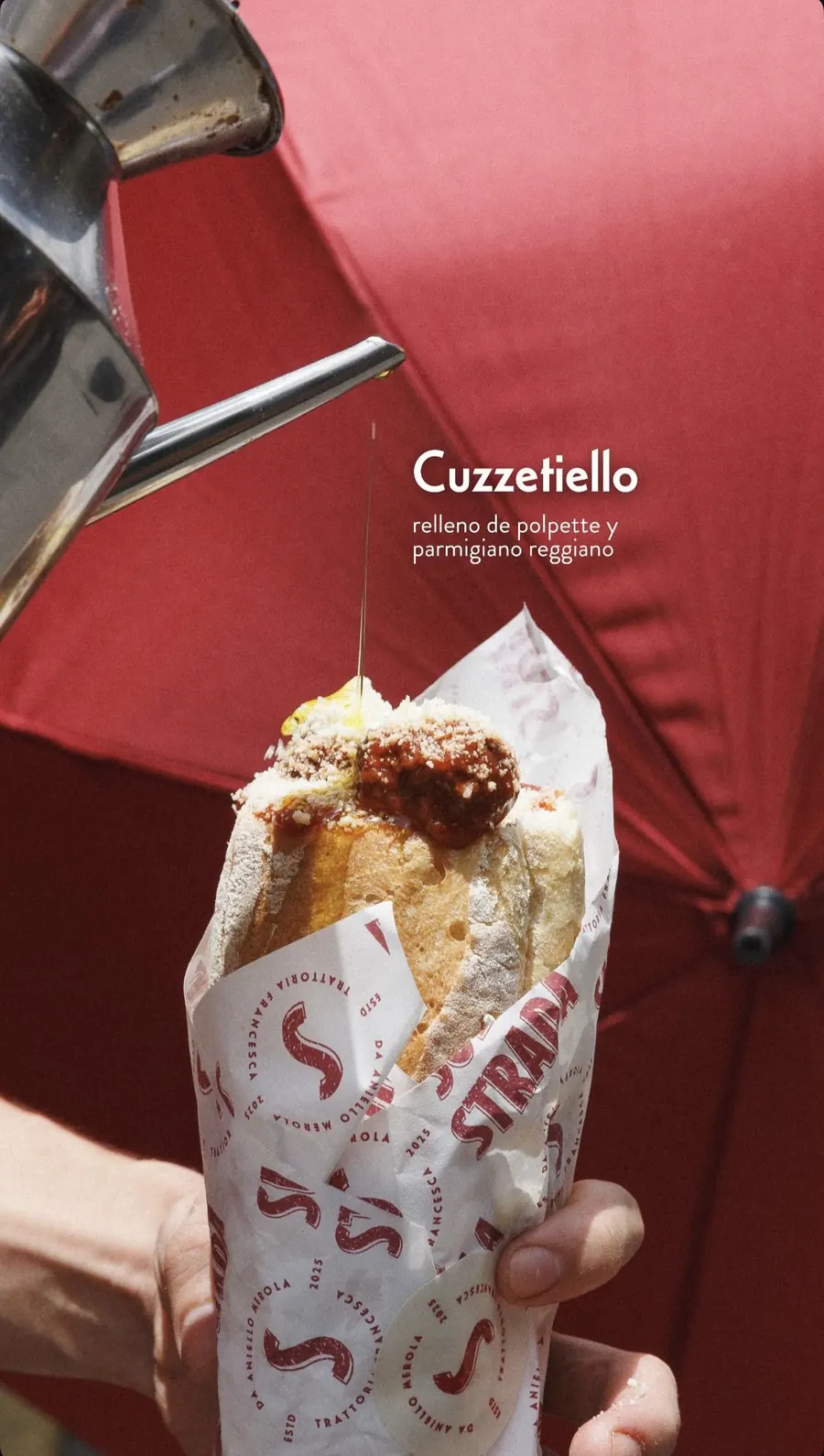 Mano sosteniendo un cuzzetiello envuelto en papel de Sulla Strada mientras una aceitera vierte aceite sobre las polpette con parmigiano, frente a un paraguas rojo