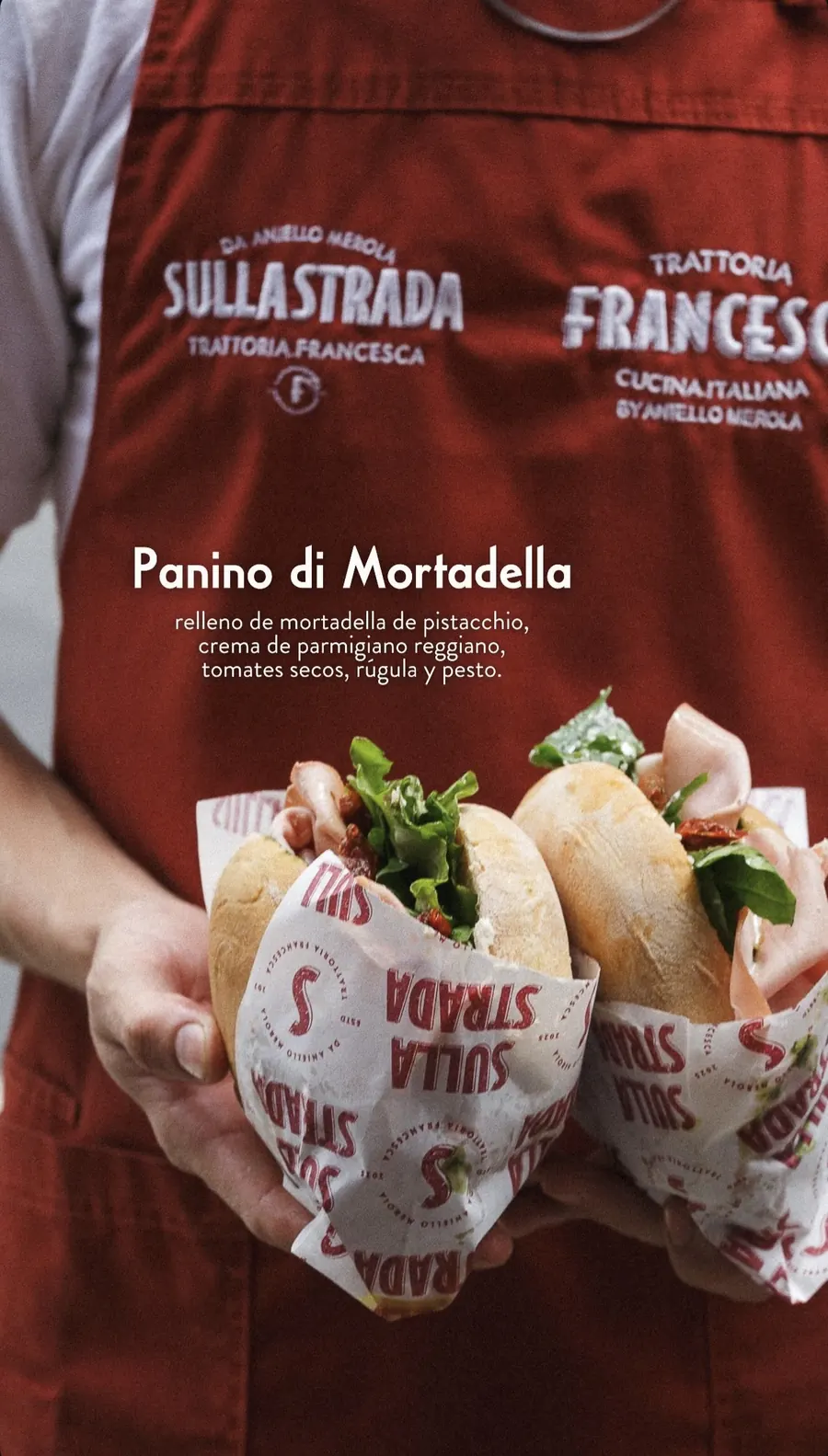 Cocinero con delantal rojo bordado de Sulla Strada sosteniendo dos panini de mortadella con rúgula, envueltos en papel de la casa