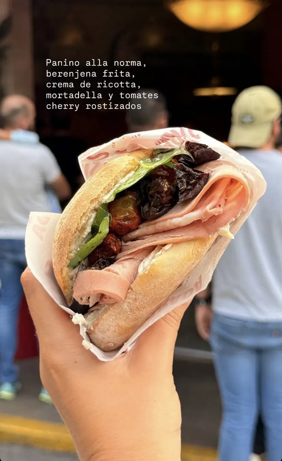 Panino alla norma con mortadella, berenjena y tomates cherry rostizados, sostenido en la acera con gente haciendo fila detrás