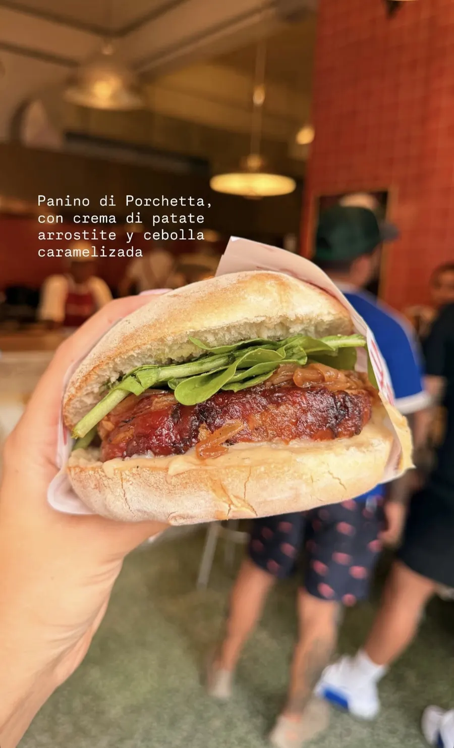 Panino de porchetta con cebolla caramelizada y hojas verdes, sostenido dentro del local con la cocina de fondo