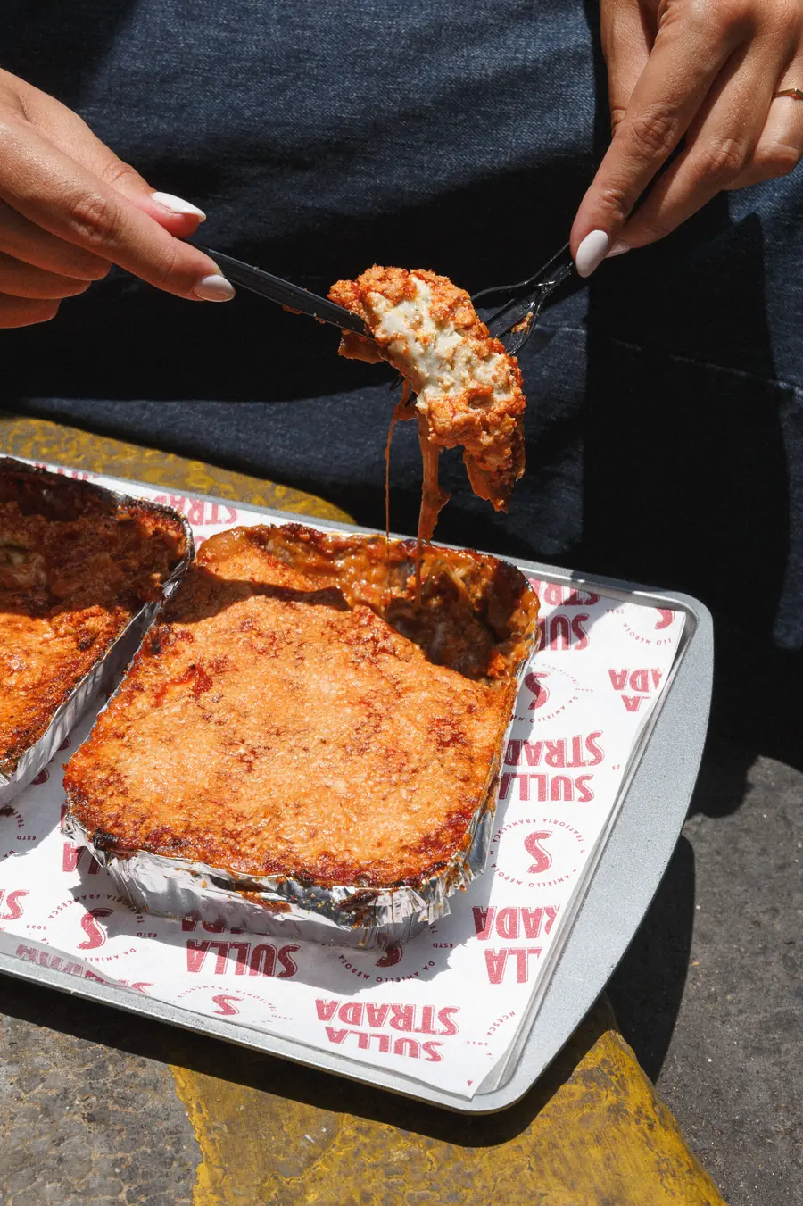 Porción de parmigiana gratinada levantada con una espátula desde su bandeja de aluminio, sobre papel impreso con el logo de Sulla Strada