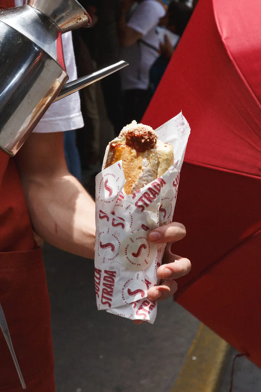 Camarero de delantal rojo sirviendo salsa sobre una pizza fritta envuelta en papel de Sulla Strada, junto a un paraguas rojo